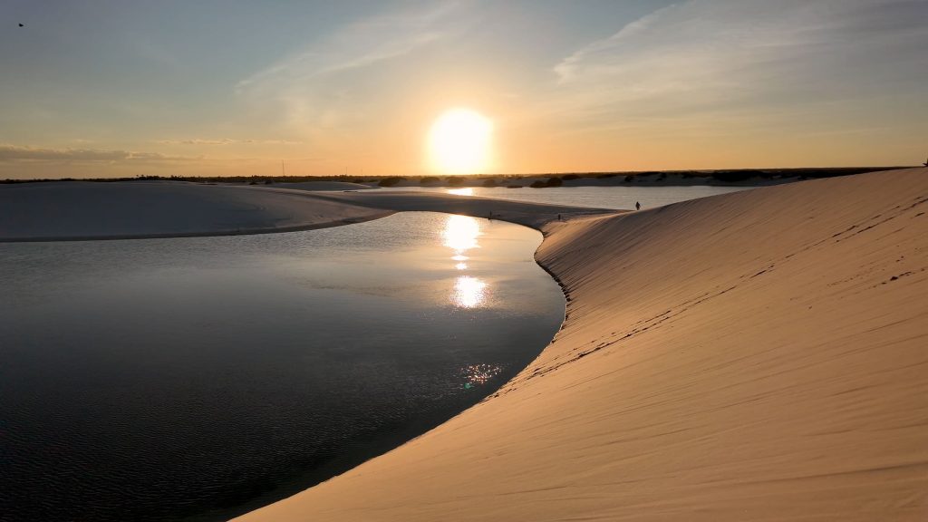 Witness a breathtaking golden hour at Lençóis Maranhenses National Park in northeast Brazil, where the setting sun casts a warm glow over the majestic dunes and tranquil lagoons, creating a mesmerizing reflection.